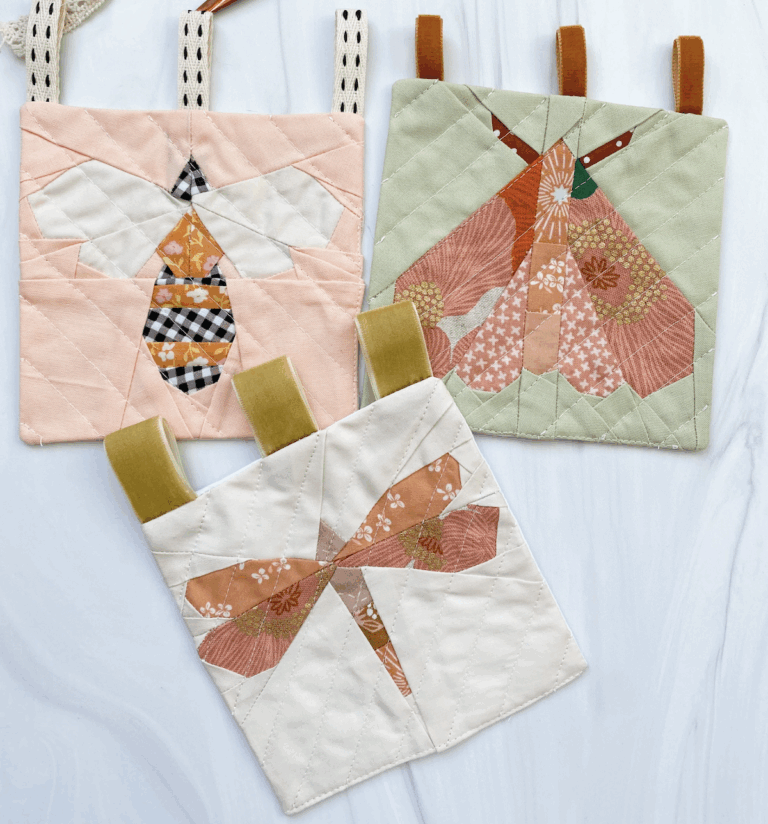 Three quilted mini panels featuring foundation paper pieced insect designs by Liza Taylor Handmade: a bee, a moth, and a dragonfly. Each design showcases soft, coordinated fabrics in shades of peach, pink, green, and cream, with hanging loops stitched along the top.